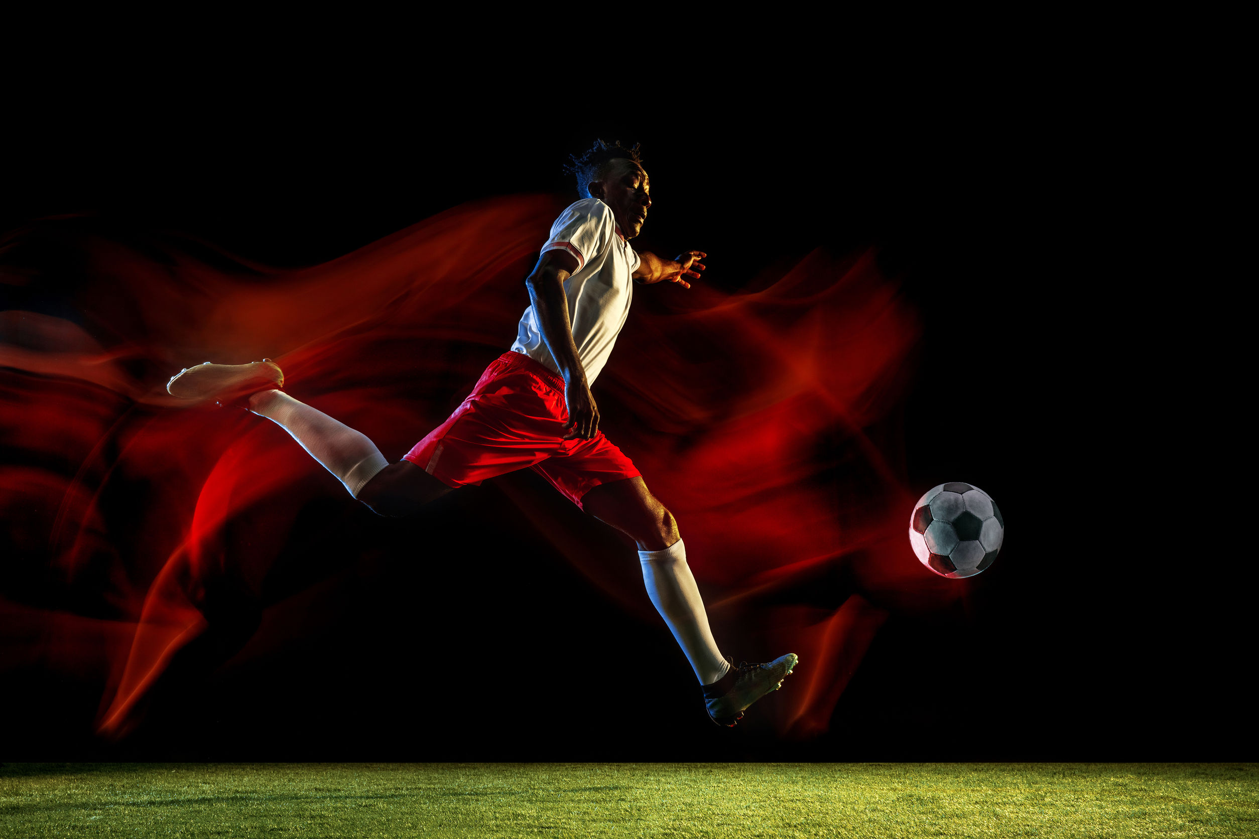 Male soccer player kicking ball on dark background in mixed light